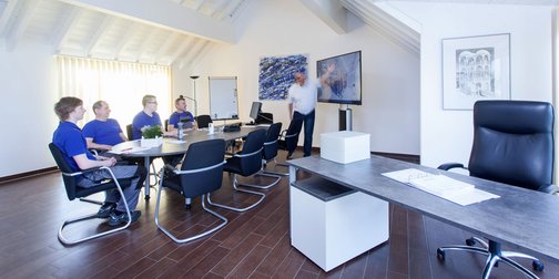 Im Konferenzraum sind fünf Männer in blauen Shirts versammelt, während einer von ihnen gestikuliert und Informationen präsentiert.