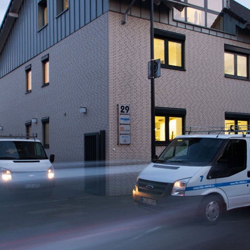 Die Fassade eines modernen Gebäudes wird von zwei Lieferwagen flankiert, während die Umgebung in sanftes Licht getaucht ist.