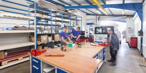 Die Szene zeigt eine gut organisierte Werkstatt mit Regalen voller Materialien und drei Männern, die an einem Projekt arbeiten.