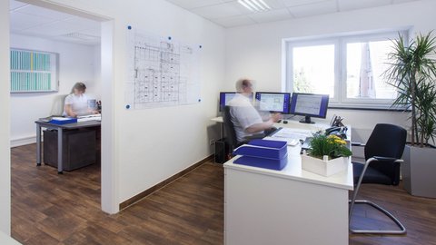 Ein modernes Büro mit zwei Arbeitsplätzen, einer Person, die am Schreibtisch arbeitet, und einer anderen, die an einem Computer sitzt.