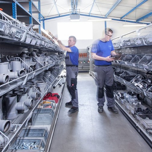 Die Regale sind gefüllt mit grauen Rohren und Zubehör, während zwei Männer konzentriert nach passenden Teilen suchen.