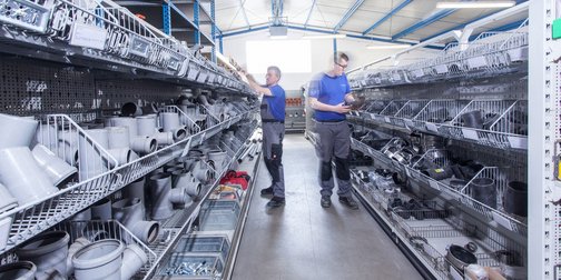 Die Regale sind gefüllt mit grauen Rohren und Zubehör, während zwei Männer konzentriert nach passenden Teilen suchen.