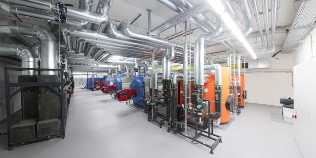 Der Raum zeigt eine Vielzahl von Heizkesseln in Blau, Rot und Orange, umgeben von glänzenden Metallrohren und einer hellen Beleuchtung.