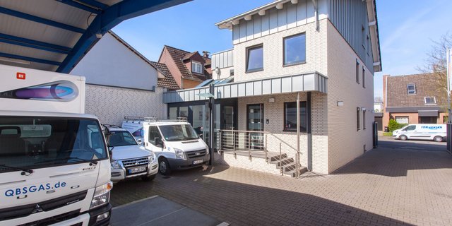 Die Fassade des Unternehmensgebäudes zeigt helle Ziegelsteine und große Fenster, während Lieferwagen im Vordergrund parken.