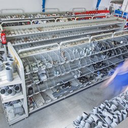 Regale mit verschiedenen Rohrarten und Zubehörteilen füllen den Raum, während zwei Mitarbeiter in blauen Shirts aktiv sind.